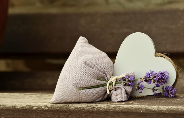 Ein helles Duftsäckchen mit einem Lavendelzweig liegt auf einer hölzernen Unterlage und ist verziert mit einem weißen Holzherz.