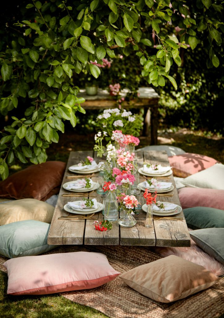 Auf einem niedrigen Holztisch mit Sitzkissen stehen Geranien und Duftgeranien in Glasbehältern. Gedeckt ist der Tisch mit sechs Tellern, auf denen ebenfalls kleine Geranienblüten liegen.