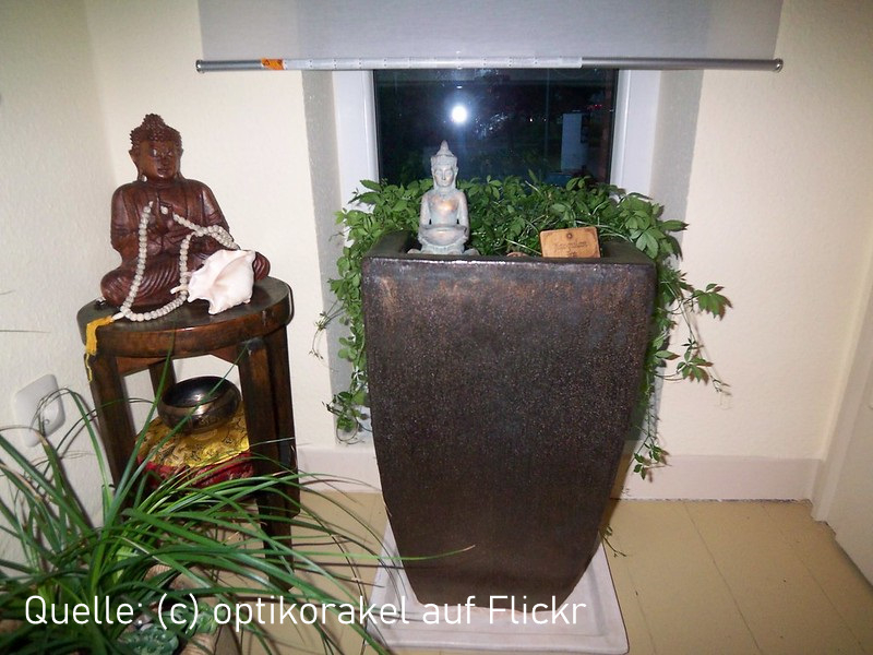 Eine asiatisch gestaltete Zimmerecke mit einem riesigen, braunen Kübel, bepflanzt mit Kraut der Unsterblichkeit und dekoriert mit einer Buddha-Figur. Daneben steht ein runder Tisch mit einer braunen Buddha Statue. Vor dem Tisch steht ein Dracanea.