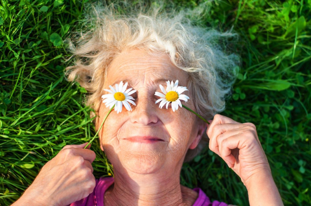 Eine glücklich lächelnde Seniorin hält sich Margeriten-Blüten vor die Augen und liegt dabei in einer grünen Wiese.