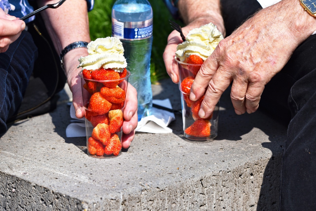 Die Hände von zwei älteren Hobbygärtnern50+, die auf einer Steinbank sitzen und frische Erdbeerbecher halten.