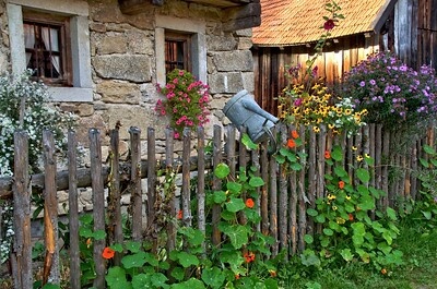 Vor einem urigen Haus steht ein bepflanzter Staketenzaun. Auf dem dem Staketenzaun hängt kopfüber eine leere eiserne Gießkanne