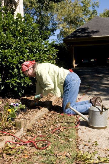 Eine Frau mit mit Jeans und Kopftuch liegt im Gartenbeet auf den Knie und pflanzt Blumen.