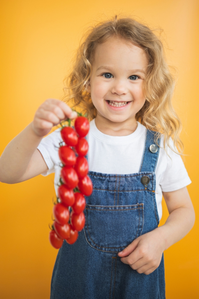 Ein kleines, blondes Mädchen mit blauer Latzhose hält lachend eine Rispe Kirschtomaten hoch.