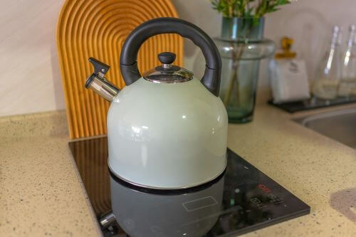 Ein weißer Wasserkessel mit schwarzem Griff steht auf einer Cerankochplatte. Im Hintergrund sieht man Küchenutensilien.