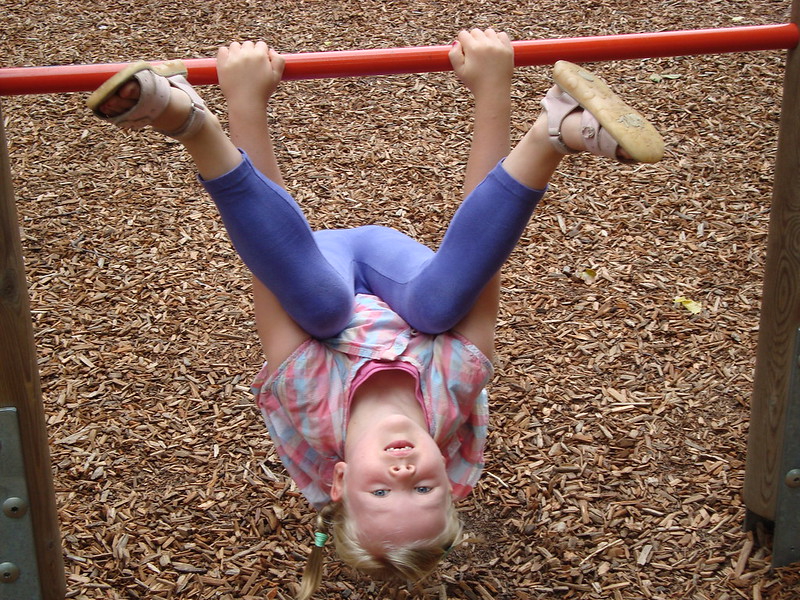 An einem Reck auf einem Spielplatz hängt ein Mädchen kopfüber. Unter dem Klettergerüst liegt eine Schicht aus Rindenmulch.