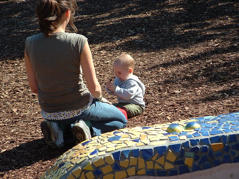 Auf einer gemulchten Fläche sitzen eine junge Mutter und ihr Kleinkind. Die Mutter kniet und das Kind sitzt. Im Vordergrund ist eine blau-gelbe Mauer zu sehen.