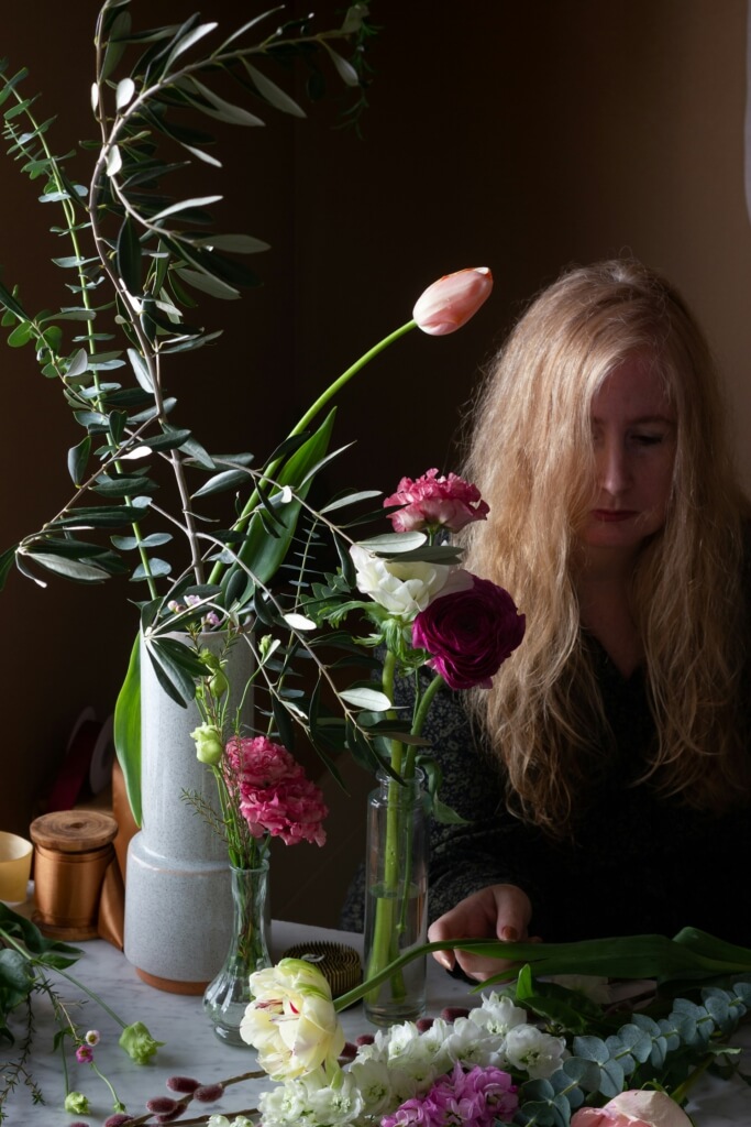 Eine junge Frau mit langen blonden Haaren sitzt an einem Tisch mit mit Blütenstängeln von Rittersporn und anderen Sommerblumen. Neben ihr stehen zwei Vasen.