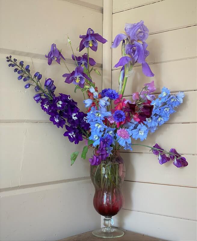 In einer Ecke aus weißen Holzvertäfelungen steht eine Bodenvase mit hellblauen und violetten Rittersporn Blütenstängeln und weiteren Sommerblumen.
