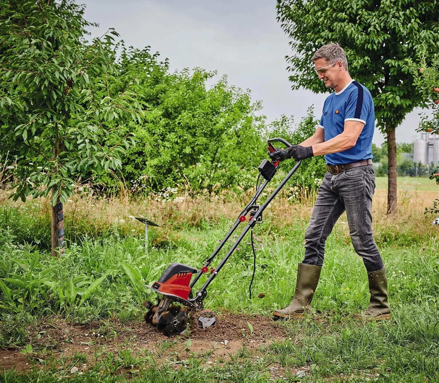 In einem stark verunkrauteten Beet schiebt ein Mann die Einhell Elektro-Boden Bodenhacke vor sich her, die die Erde auflockert.