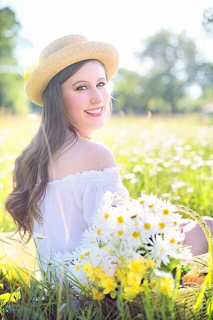 Eine junge Frau mit Sonnenhut dreht sich lächelnd zur Kamera und sitzt in einer Blumenwiese.