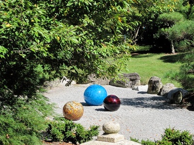 In einem Zengarten mit einer Rasenfläche im Hintergrund, Kies als Untergrund, Konifieren und Findlingen liegen drei große farbige Steinkugeln in hellblau, dunkelrot und hellbraun.
