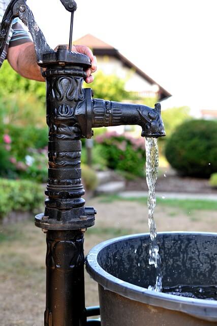 An einer schwarzen Gartenpumpe pumpt ein Mann Wasser in einen schwarzen Bottich.