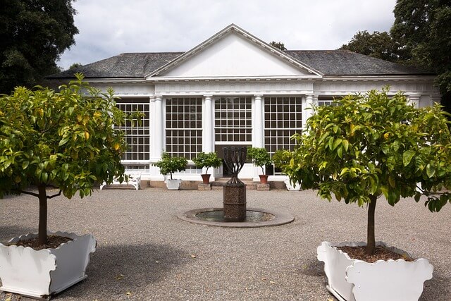 Vor einer großen, weißen Orangerie stehen mehrere Zitronenbäume in weißen Kübeln auf einem weitläufigen Vorplatz.