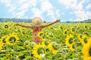 Eine Frau in Sommerkleidung steht mit ausgebreiteten Armen in einem Sonnenblumenfeld.